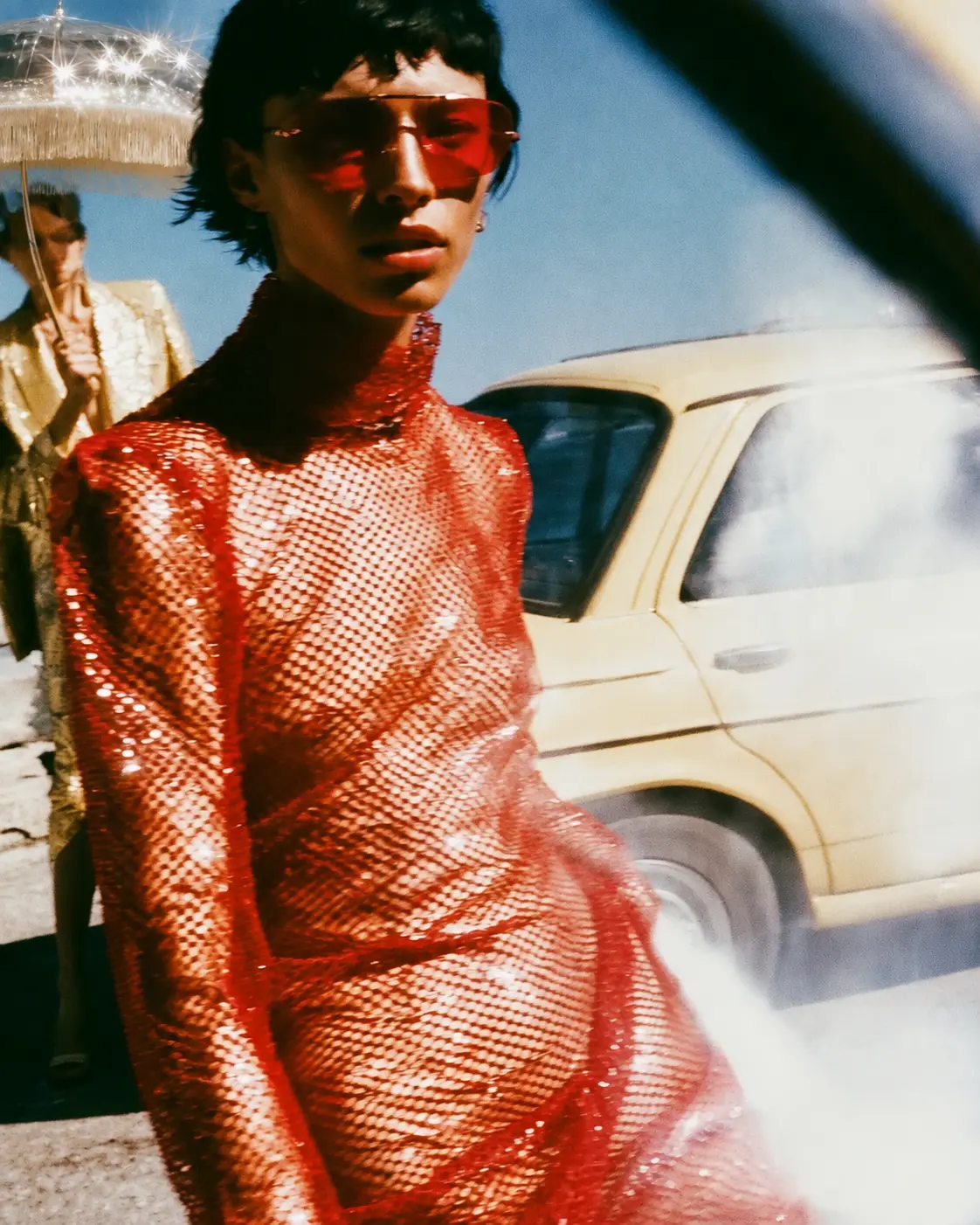 Woman in a red mesh dress and red sunglasses beside a cream car, another figure holding a fringed umbrella behind