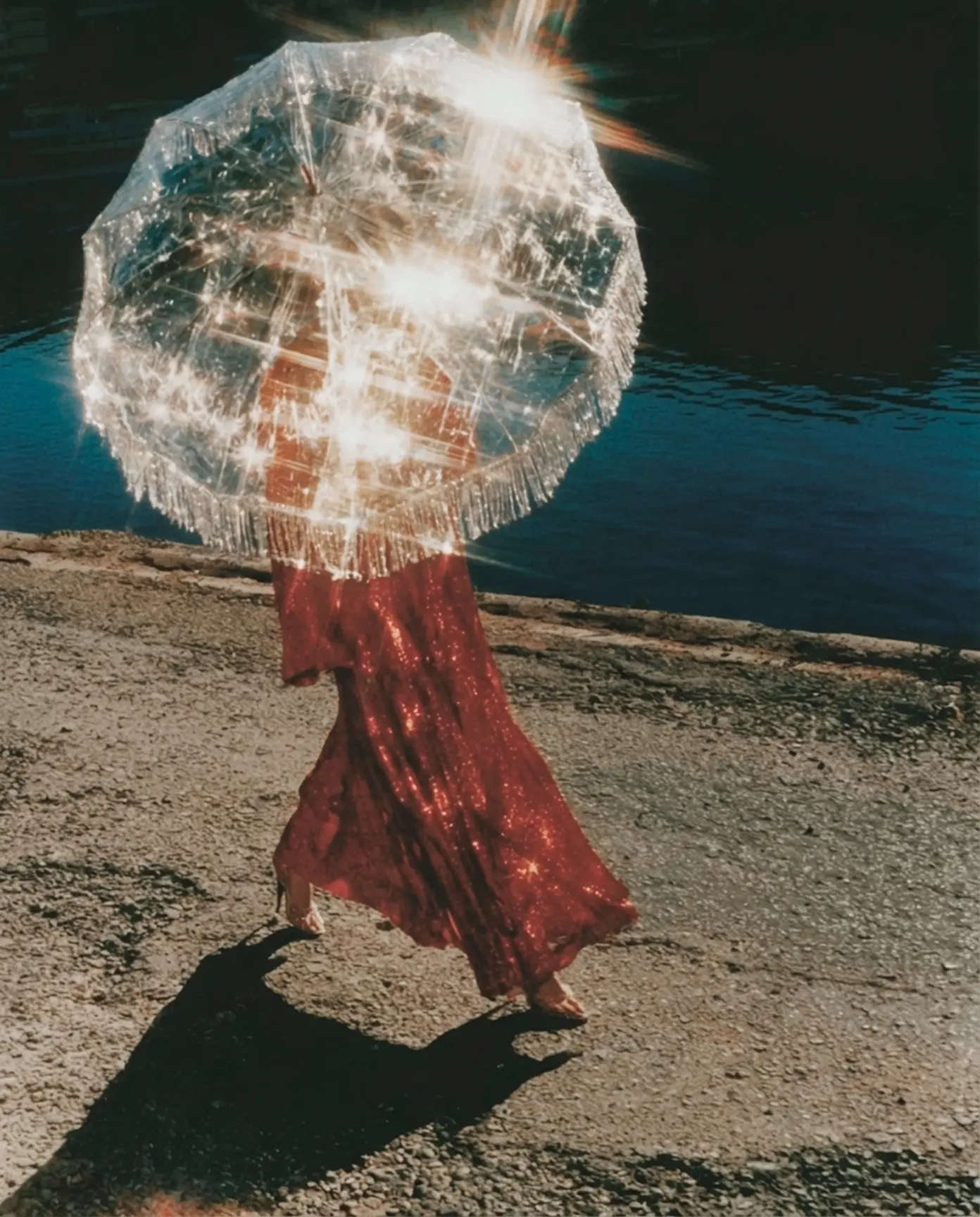 Woman in a red sequin skirt walking under a clear fringed umbrella flaring in the sun by dark water