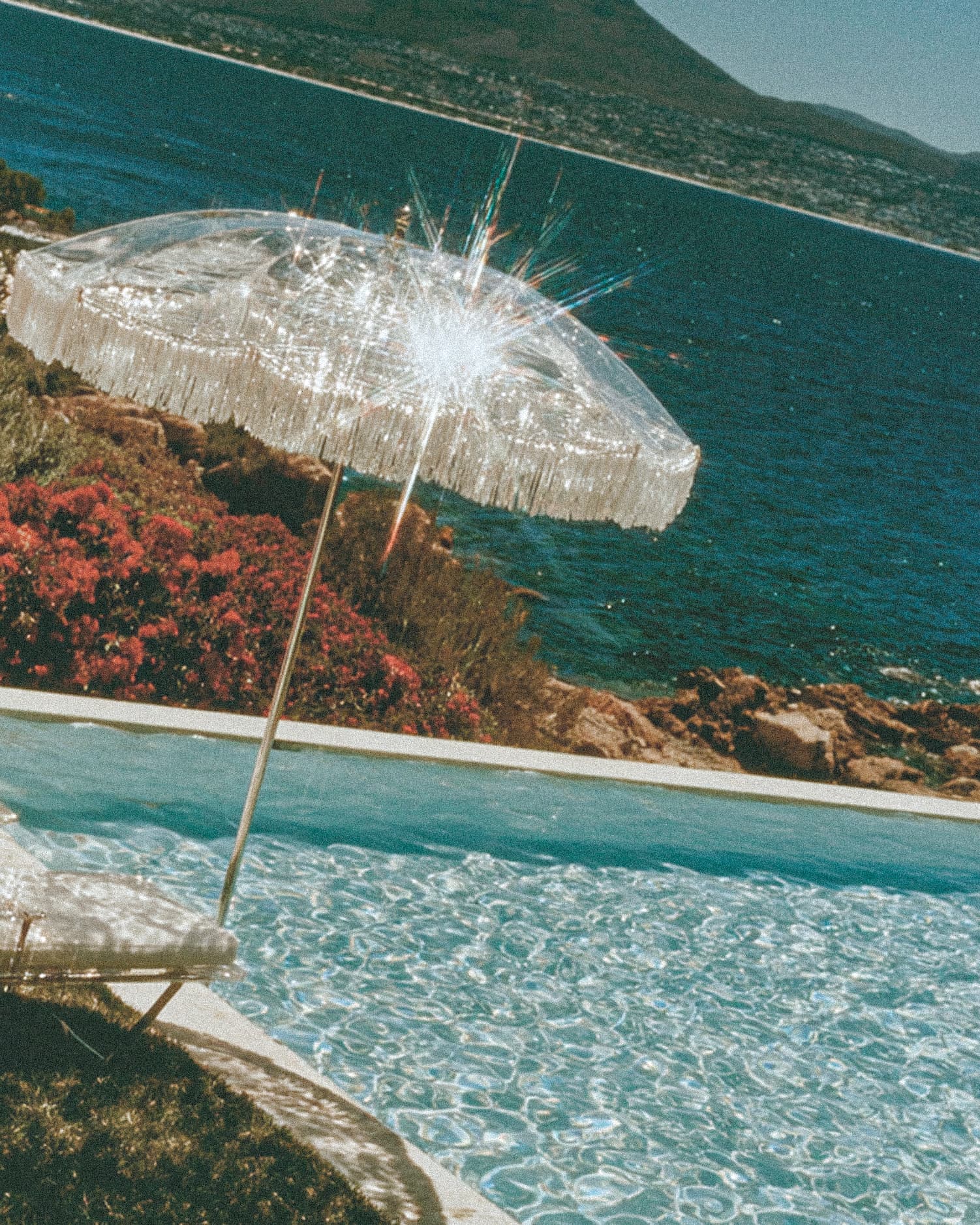 Clear fringed umbrella sparkling in the sun above a pool, with red flowers, rocky coast and blue sea behind