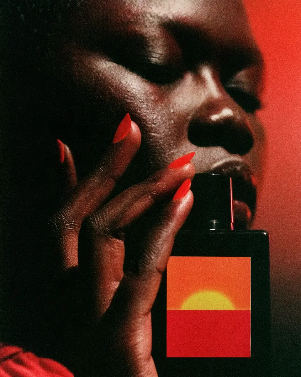 Model with red nails holding a black fragrance bottle with a sunset label to her cheek