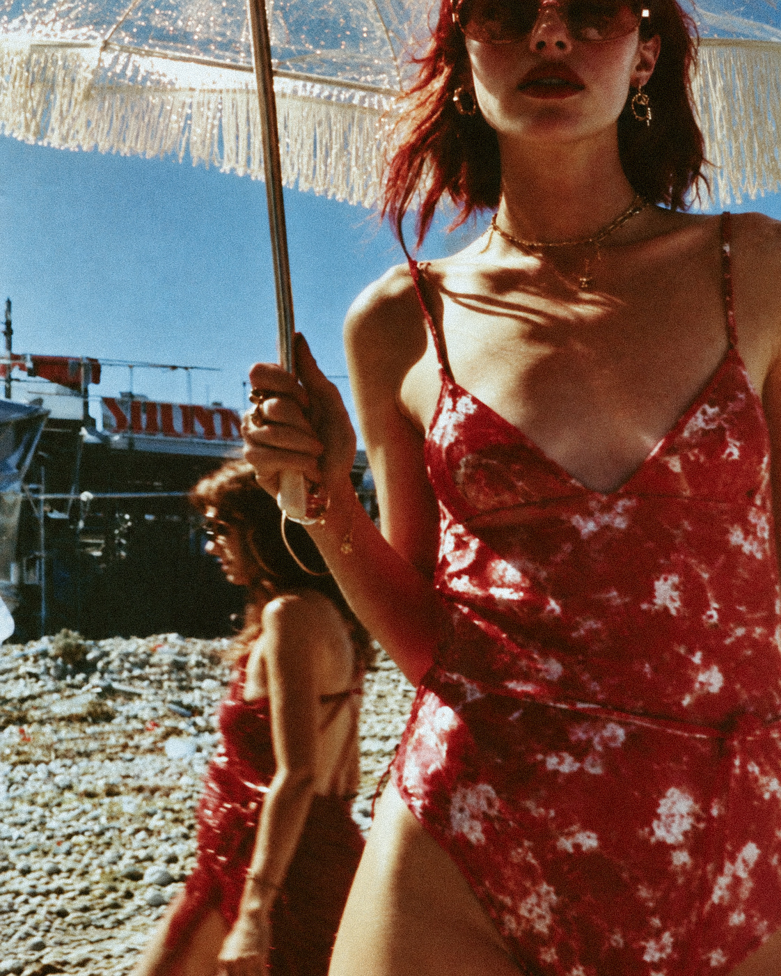Woman in a red floral dress holding a fringed umbrella near a boat on a pebble beach