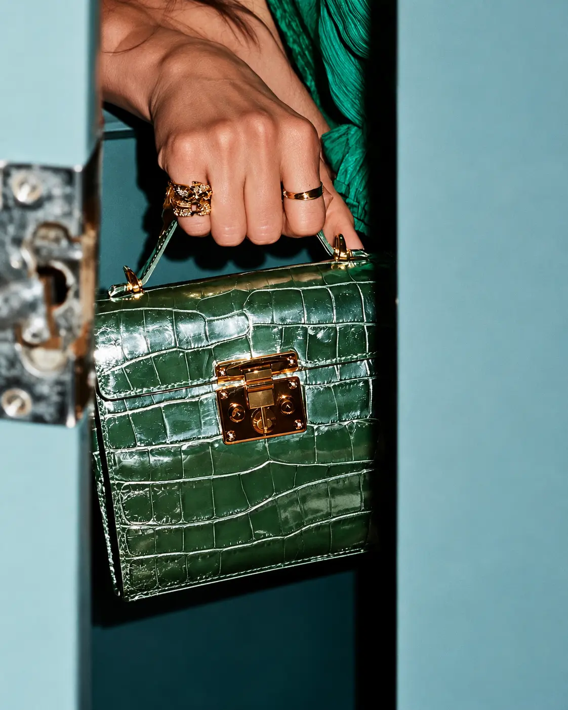 Hand with gold rings holding a green croc bag in a blue doorway
